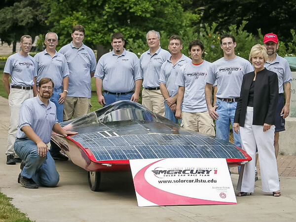 A group of eleven people standing behind a solar car covered in panels.