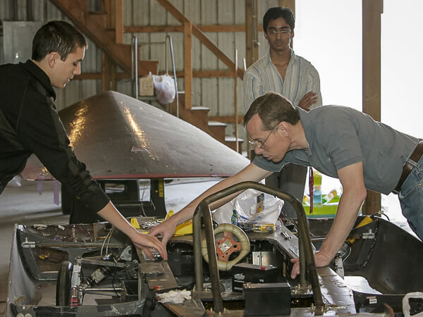Three individuals examine a solar car in a workshop setting.
