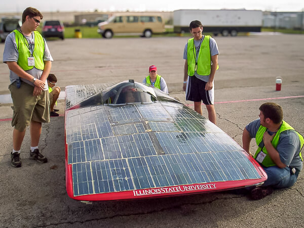 Solar-powered vehicle with solar panels and "Illinois State University" written on it, surrounded by five people in neon vests.