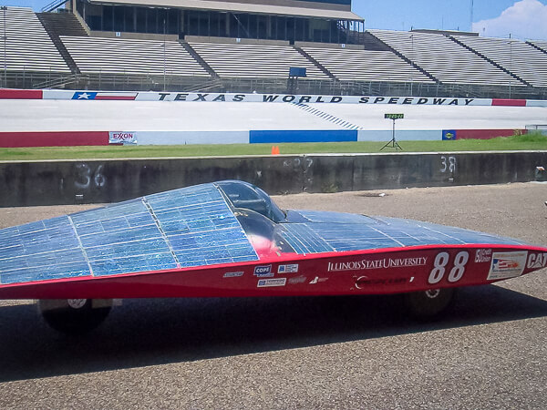 A solar car with a red frame and dark blue solar panels parked in front of the Texas World Speedway grandstand.