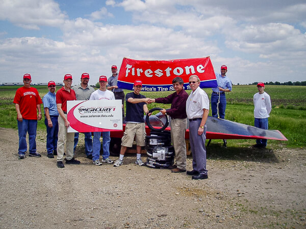 A group of people stand in front of a solar car with a red Firestone banner behind them, on a dirt road.