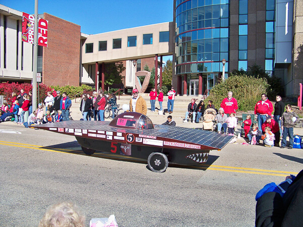 A solar-powered car with a flat solar panel body and a bubble canopy in a parade, surrounded by spectators next to a campus building.