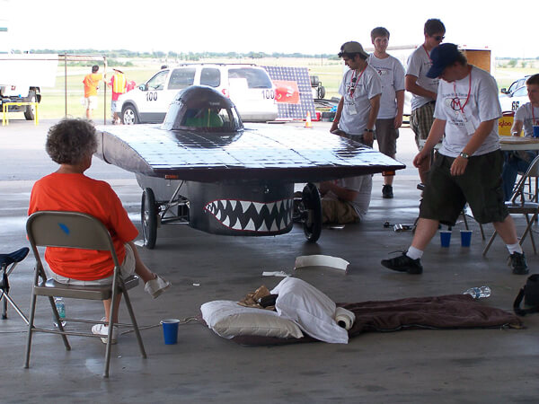 Several people gather around a solar-powered car with a shark mouth design inside an open hangar.