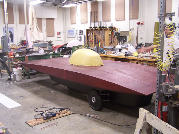 A maroon, oval-shaped solar car with a yellow dome in a workshop.