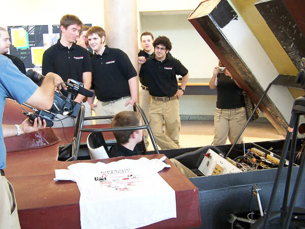 A group of people in black shirts and beige pants examine an open solar car project indoors, with one person filming and another sitting inside.