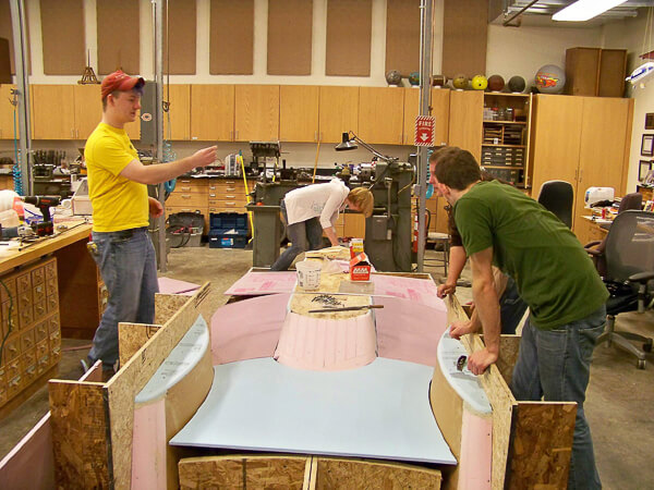 Three people working on a foam and wood structure in a workshop.