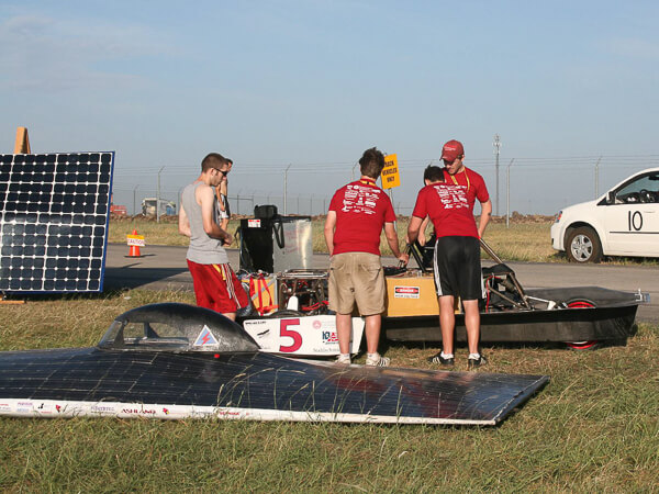 Four people examine a solar-powered vehicle on a grassy field near a solar panel and a white van.