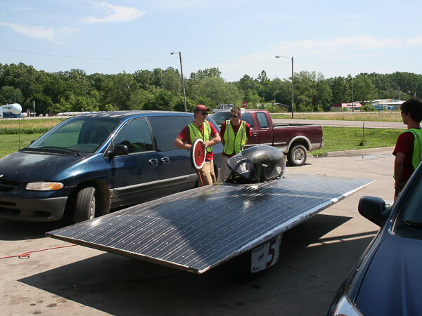 Solar-powered vehicle with solar panels on a paved area, surrounded by three people in high-visibility vests.