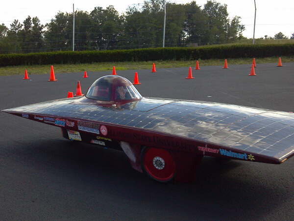 Solar-powered car with "Illinois State University" and sponsor logos, on a paved area with traffic cones in the background.