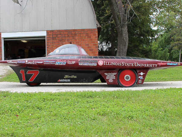 Black and red solar car with number 17 and sponsor logos, including Illinois State University, parked on a gravel path.
