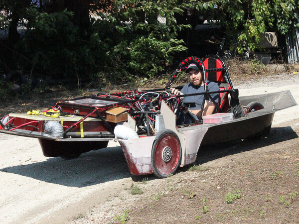 A man seated in a homemade solar car with a flat, metallic body and exposed framework.