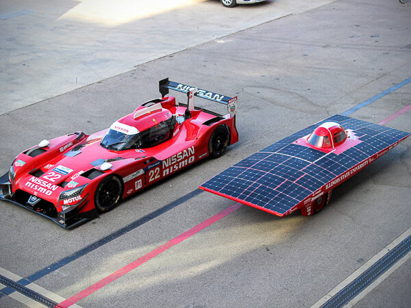 A red race car and a solar-powered vehicle parked side by side in a parking area.