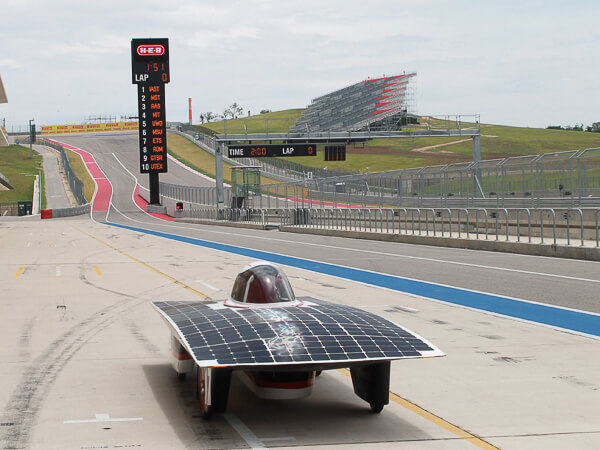 A solar-powered car on a race track with a scoreboard and grandstands in the background.
