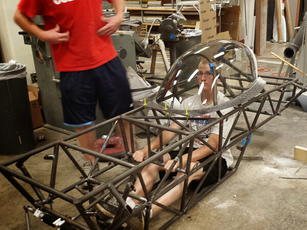 A person inside a skeletal vehicle frame with a clear canopy in a workshop.