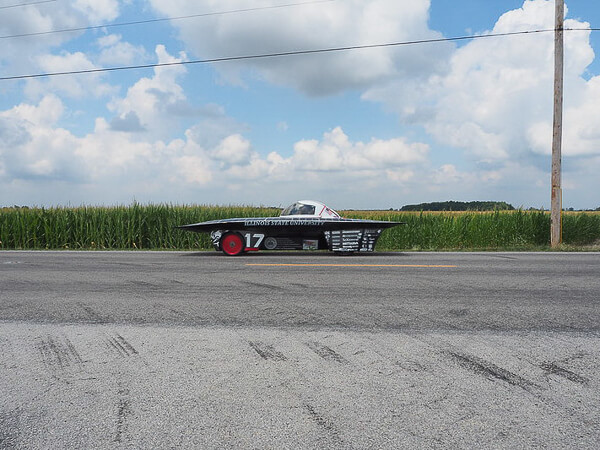 A solar car with the number 17 drives on a road beside a green cornfield under a cloudy sky.