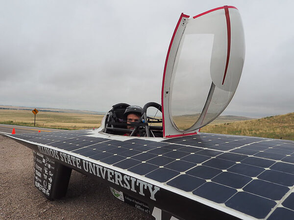 Solar vehicle with "ILLINOIS STATE UNIVERSITY" on the side, features an open canopy with a person inside, set in a rural landscape.