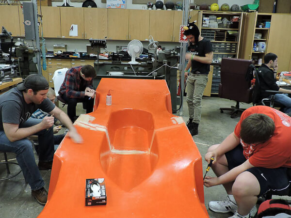 Several people work on a large, orange base in a workshop.