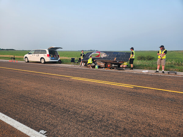 A solar-powered car with people in safety vests on the side of a rural road, next to a white minivan.