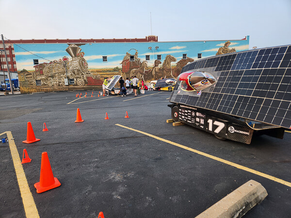 A solar-powered vehicle with a number "17" parked on a lot with a train mural in the background. Orange cones create a path nearby.