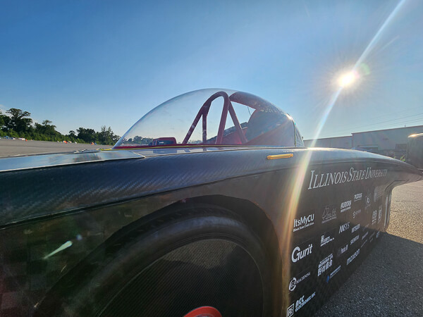 Solar car with aerodynamic design and sponsor logos on a sunlit road.